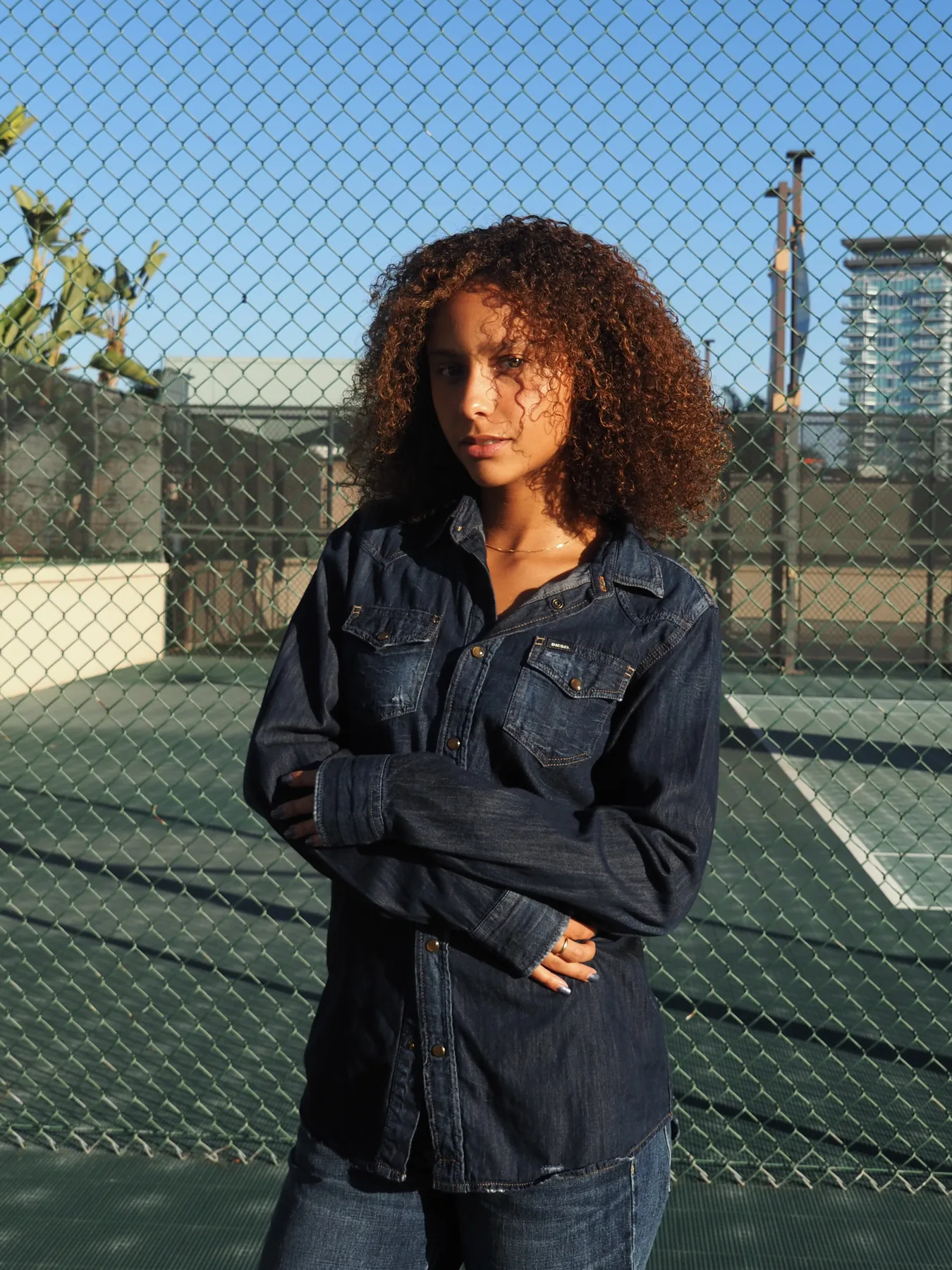 Sophia Coon in a dark denim shirt, arms folded, standing against the chain-link fence of a tennis court